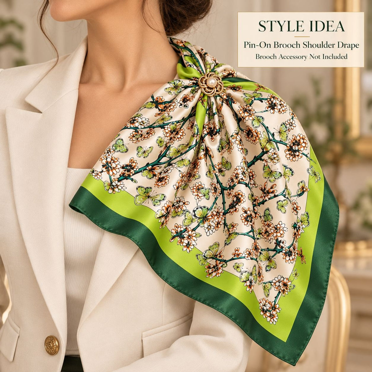 A woman wearing a cream-colored satin scarf with a floral branch print, green border, and a gold-tone brooch at the collarbone, styled over a white blazer. A text box in the top right reads 'STYLE IDEA Pin-On Shoulder Drape Brooch Accessory Not Included'.