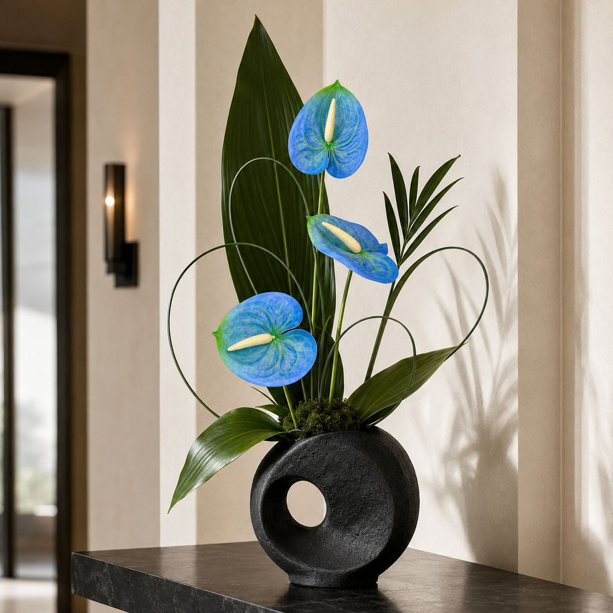 A floral arrangement of three blue anthuriums with green leaves in a round black ceramic vase, displayed on a dark table against a beige wall.
