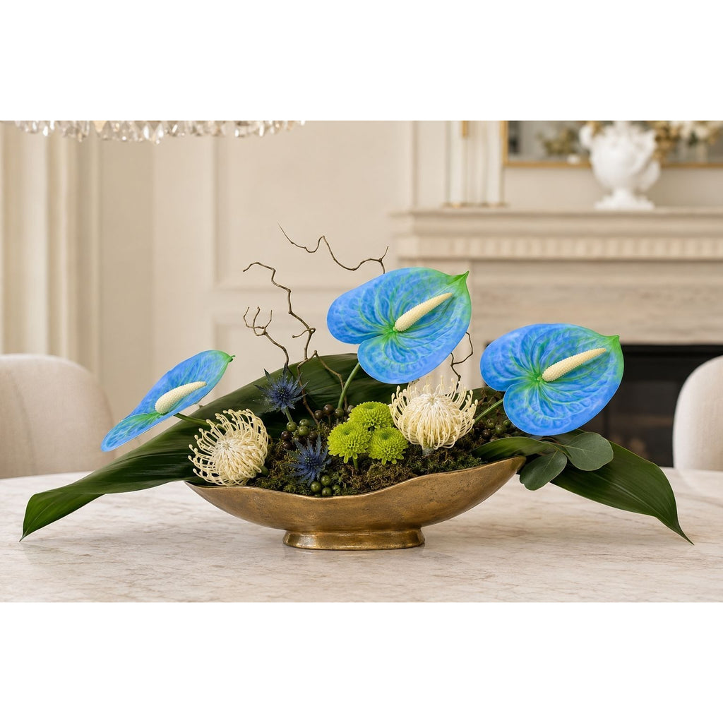 A floral arrangement in a gold bowl centerpiece, featuring vibrant blue anthuriums, green foliage, and textured cream and blue flowers, displayed on a table in a softly lit indoor setting.