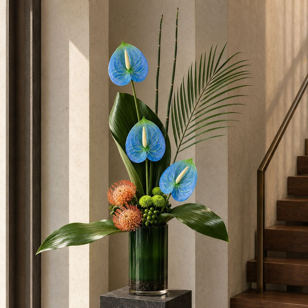 A floral arrangement in a dark green glass vase placed on a dark pedestal, set indoors beside a staircase. The arrangement features three bright blue anthurium flowers, large green leaves, palm fronds, and two round orange-brown berries.