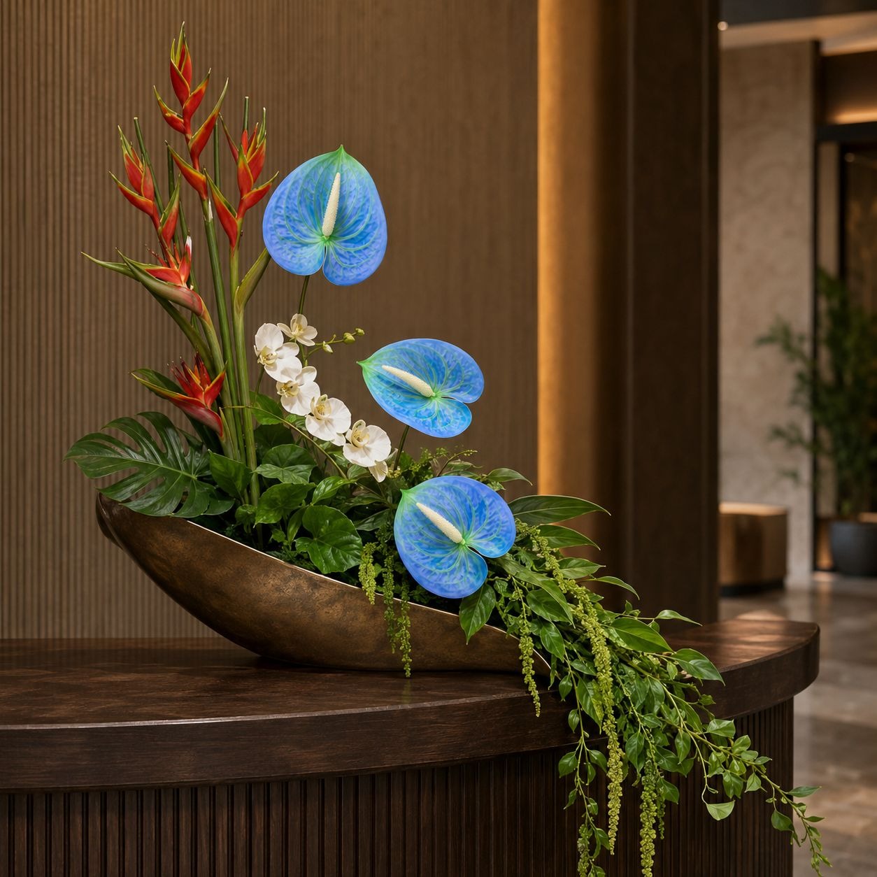 A decorative floral arrangement in a curved bronze bowl, featuring three bright blue anthuriums, red bird of paradise flowers, small white orchids, and trailing green foliage, displayed on a dark wooden counter in an indoor setting.