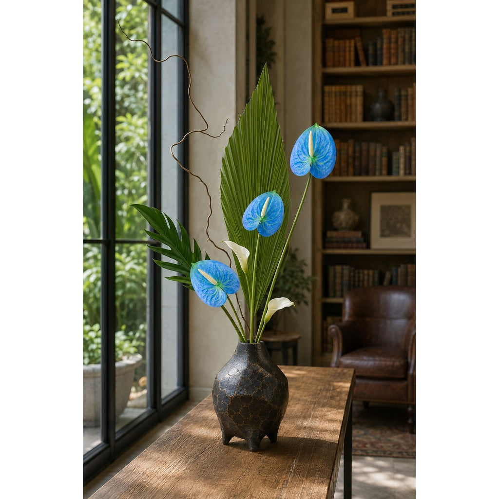 A dark textured ceramic vase holding a tall green palm frond and three bright blue anthurium flowers, placed on a wooden table in a sunlit room with a window and bookshelves in the background.