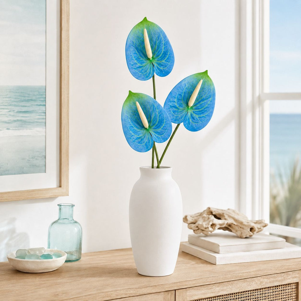 Three blue anthurium flowers with green and white tips arranged in a tall white ceramic vase on a light wood console table, with a window, framed ocean print, and small decorative objects in the background.