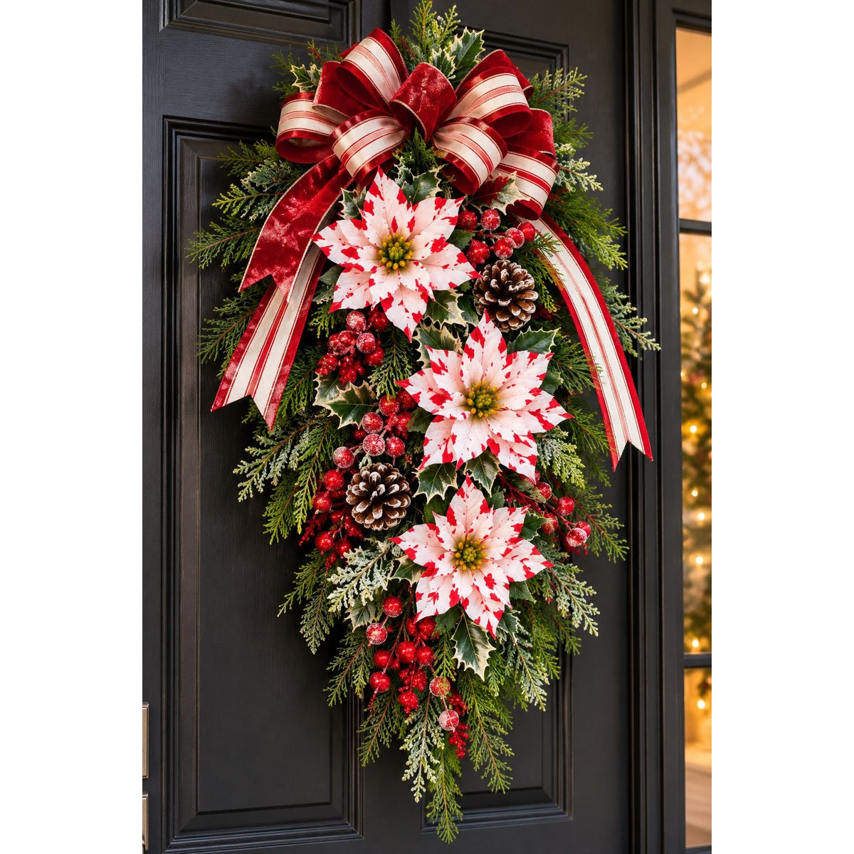 A festive Christmas wreath hanging on a black front door, featuring red and white poinsettias, green holly, pinecones, evergreen branches, and a large red and white striped bow at the top.