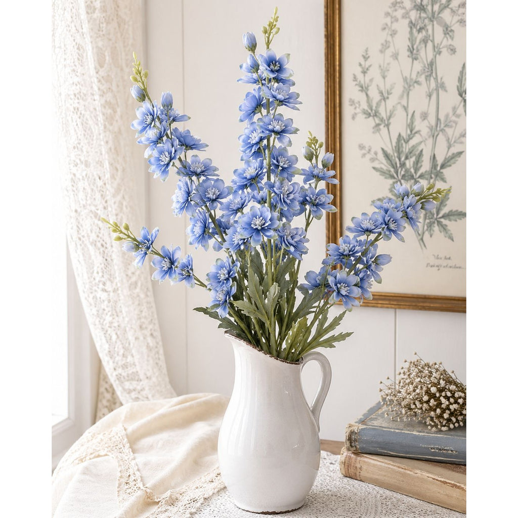 White vase with blue flowers on a surface with a framed botanical print in the background