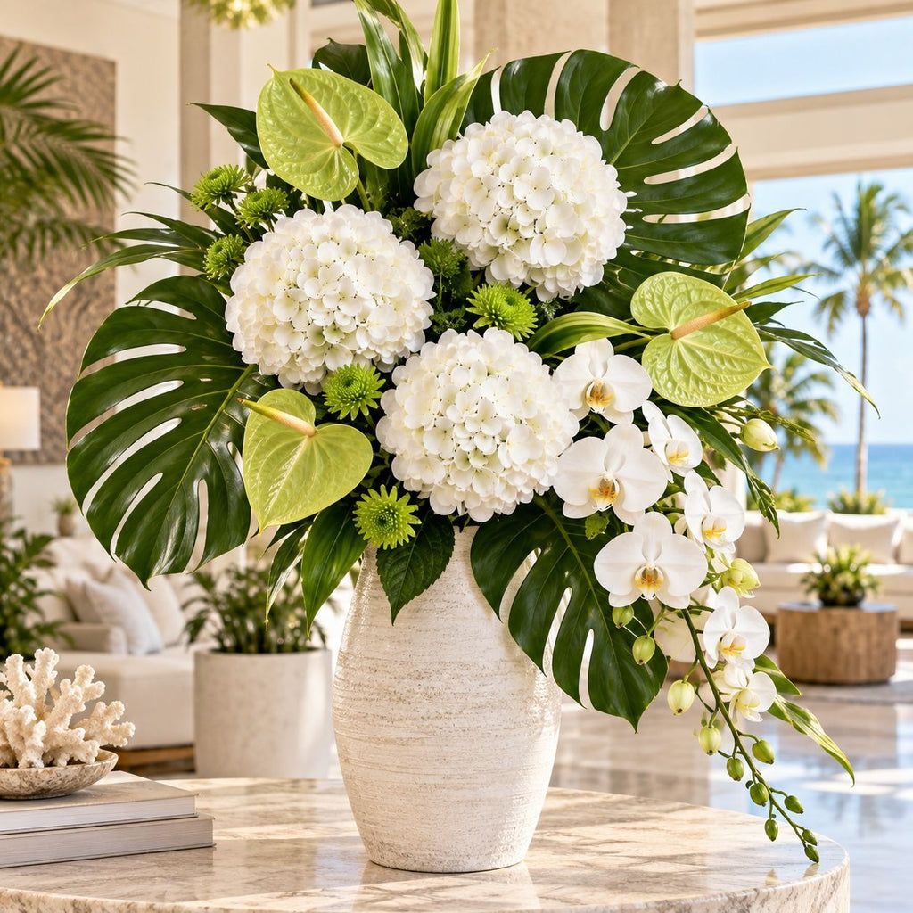 A large tropical flower arrangement in a textured cream ceramic vase, featuring white hydrangeas, green monstera leaves, anthuriums, and white orchids, displayed on a marble table in a bright, modern living room with an ocean view.