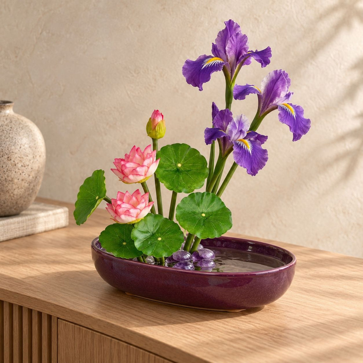 A decorative artificial flower arrangement in a glossy dark purple oval dish, featuring purple irises, pink lotus flowers, and green lily pads on a wooden table against a textured beige wall.