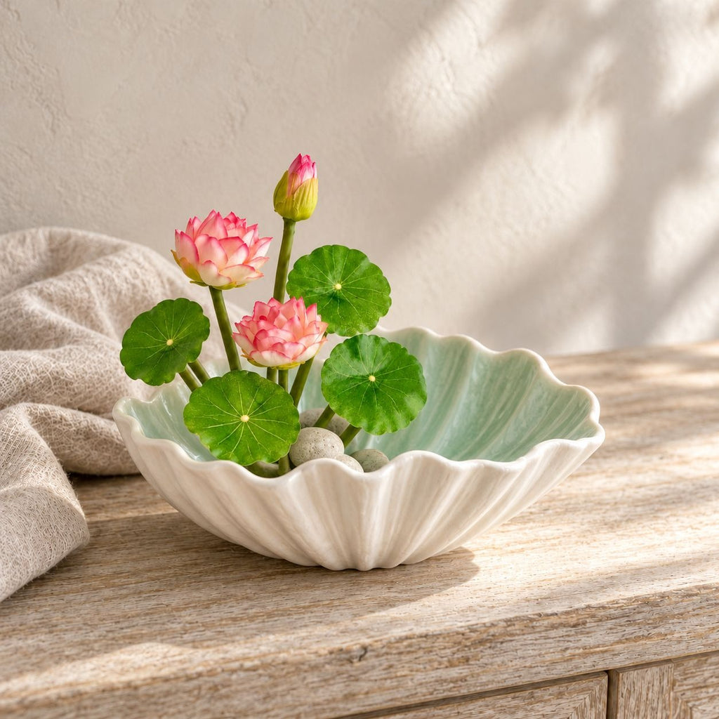 A scalloped ceramic bowl with a light green interior and ribbed cream exterior, styled with pink lotus flowers and green lily pads on a wooden table.