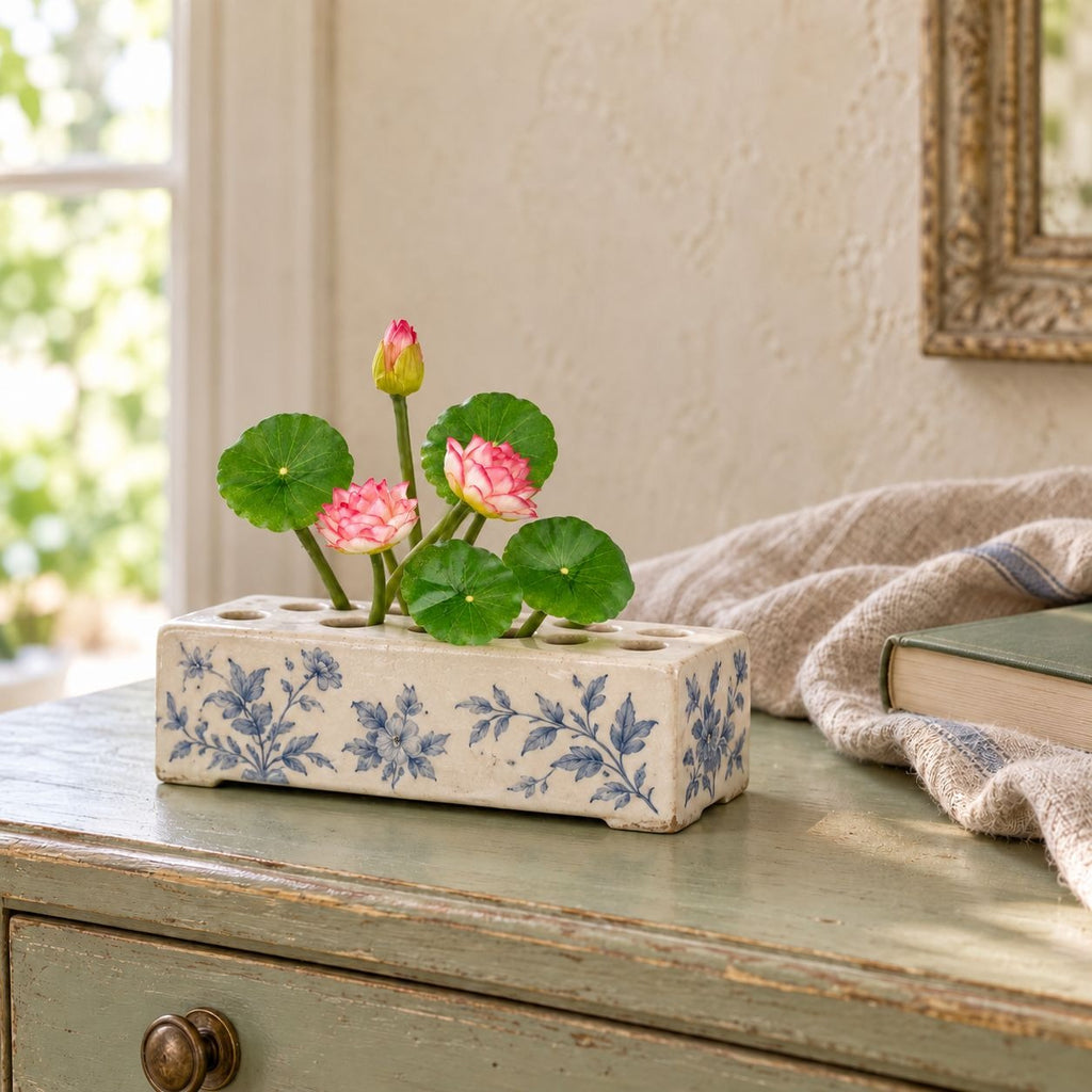 A rectangular ceramic planter with blue floral patterns sits on a distressed green wooden table, holding pink lotus flowers and green leaves in a styled interior setting.