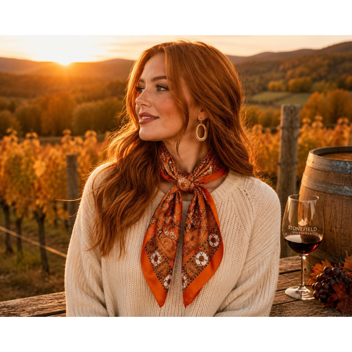 A woman with long red hair stands outdoors in a vineyard at sunset, wearing a cream knit sweater and an orange patterned neck scarf tied around her collar. A glass of red wine and a wooden barrel are visible beside her.