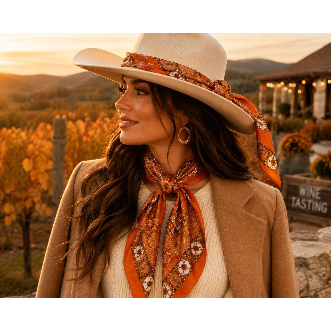 A woman outdoors in a vineyard at sunset, wearing a cream wide-brim hat with an orange paisley bandana, a cream sweater, and a tan coat, with an orange paisley neck scarf tied around her neck.