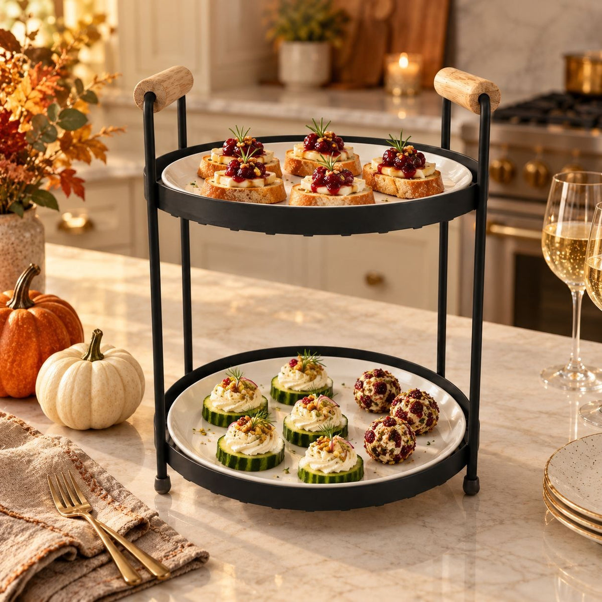 A two-tier black metal serving stand with light wood handles, holding appetizers on a marble kitchen counter. The scene includes pumpkins, fall leaves, wine glasses, and a stove in the background.