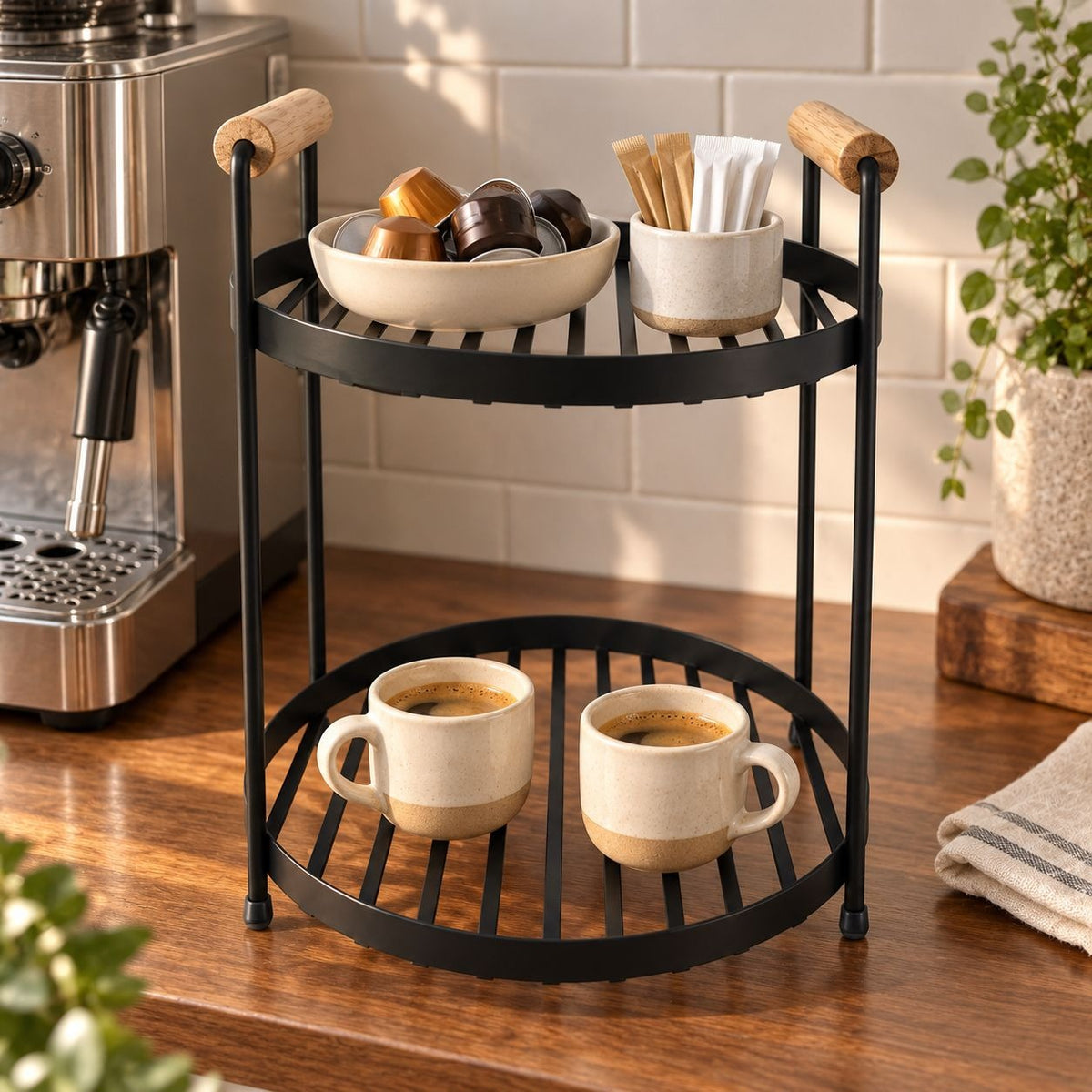 A two-tier black metal coffee station with wooden handles, styled on a wooden countertop with ceramic mugs, coffee pods, and a coffee machine in the background.