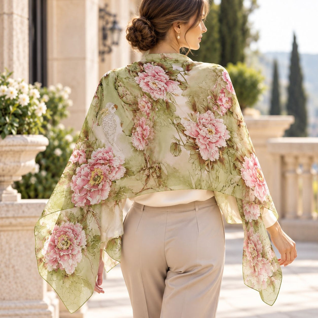 A woman viewed from behind wearing a sheer floral shawl with large pink peony-style flowers on an olive green background, paired with light beige pants in an outdoor architectural setting.