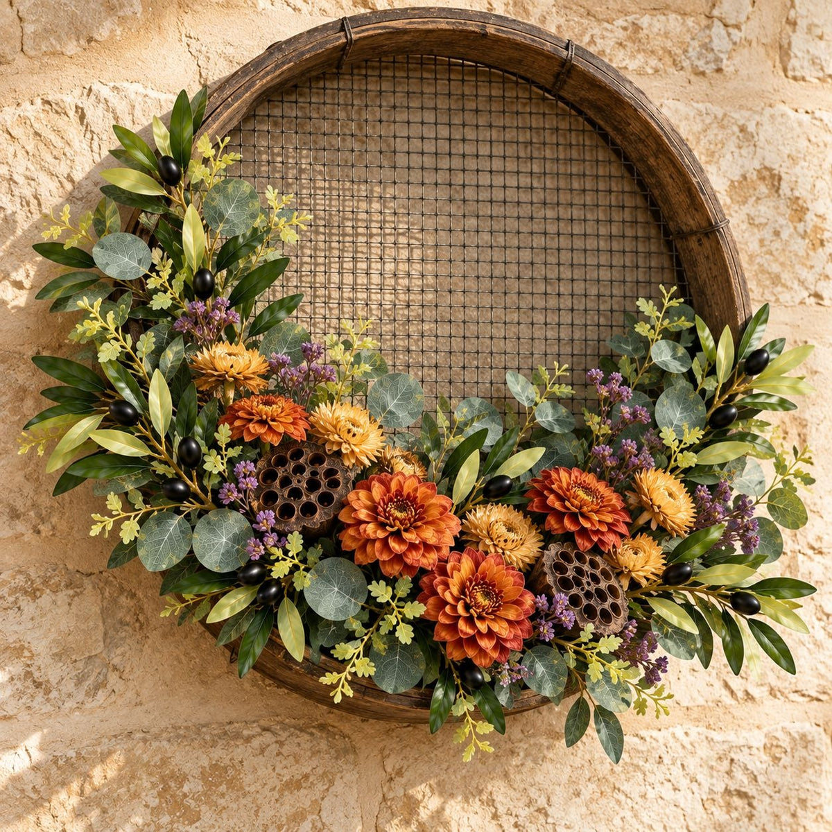 An autumn-themed rustic wreath made from a repurposed wooden barrel hoop with a wire mesh center, decorated with green eucalyptus leaves, orange and yellow dahlia flowers, purple berries, and black olives, hung on a beige stone wall.