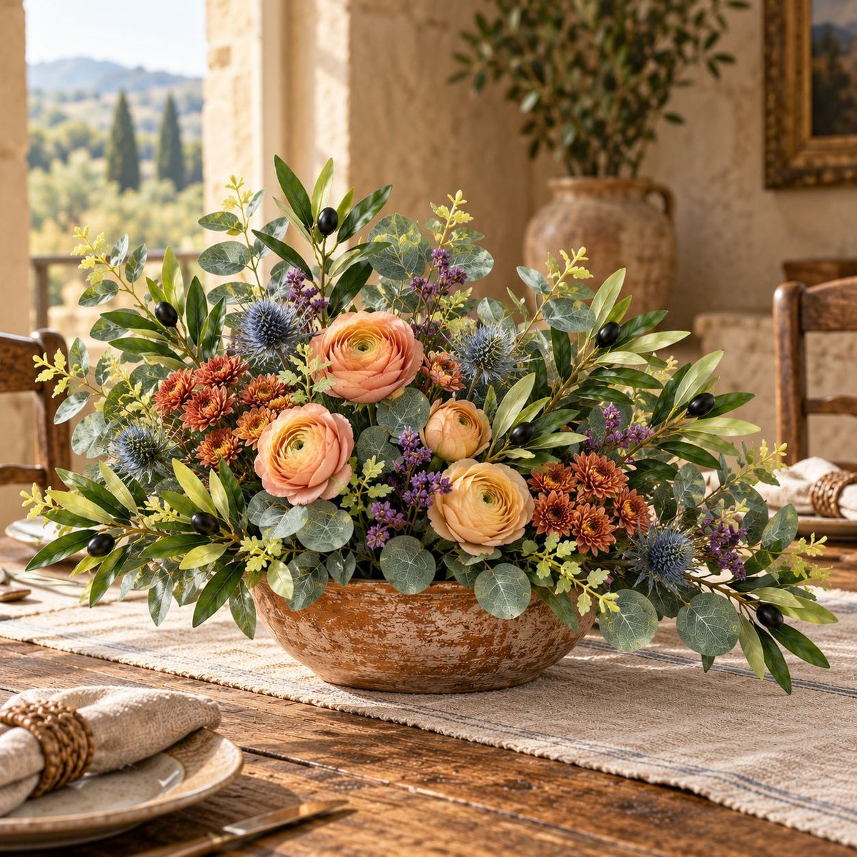A rustic floral centerpiece arrangement in a distressed brown bowl, placed on a woven runner over a wooden dining table. The arrangement features peach ranunculus, green eucalyptus, blue thistles, and orange-brown mums, with a warm, sunlit interior background.