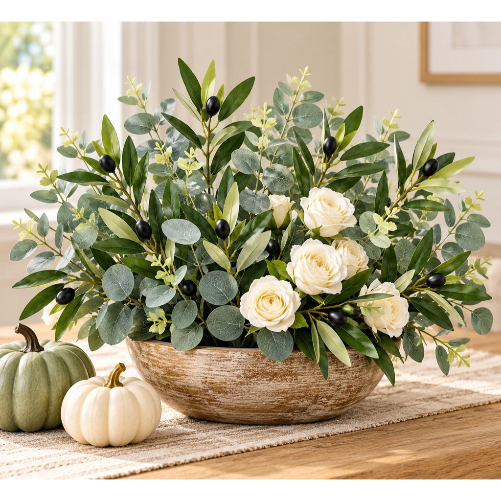 A large artificial floral arrangement in a rustic round bowl, featuring green eucalyptus leaves, white roses, and small black berries, styled on a table with two small pumpkins in a bright indoor setting.