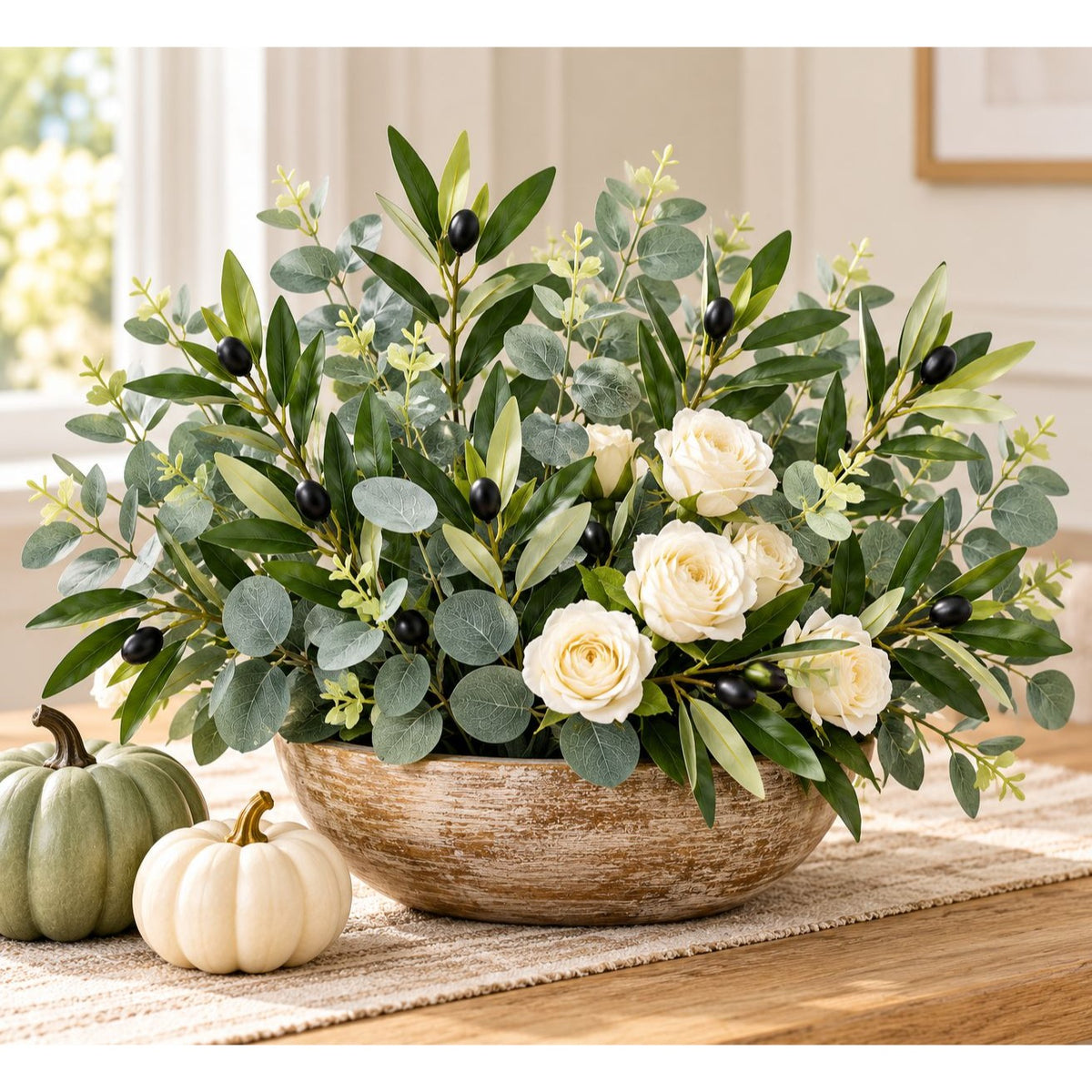 A large artificial floral arrangement in a rustic round bowl, featuring green eucalyptus leaves, white roses, and small black berries, styled on a table with two small pumpkins in a bright indoor setting.