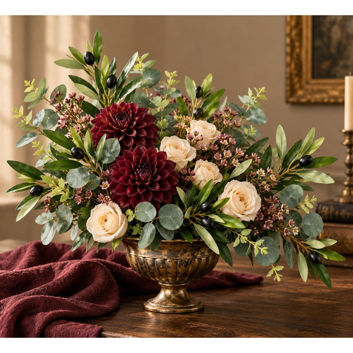 A decorative floral arrangement in an ornate gold pedestal bowl, placed on a wooden table with a draped burgundy fabric. The bouquet includes large deep burgundy dahlias, cream roses, green eucalyptus leaves, and small dark berries, set against a warm indoor background.