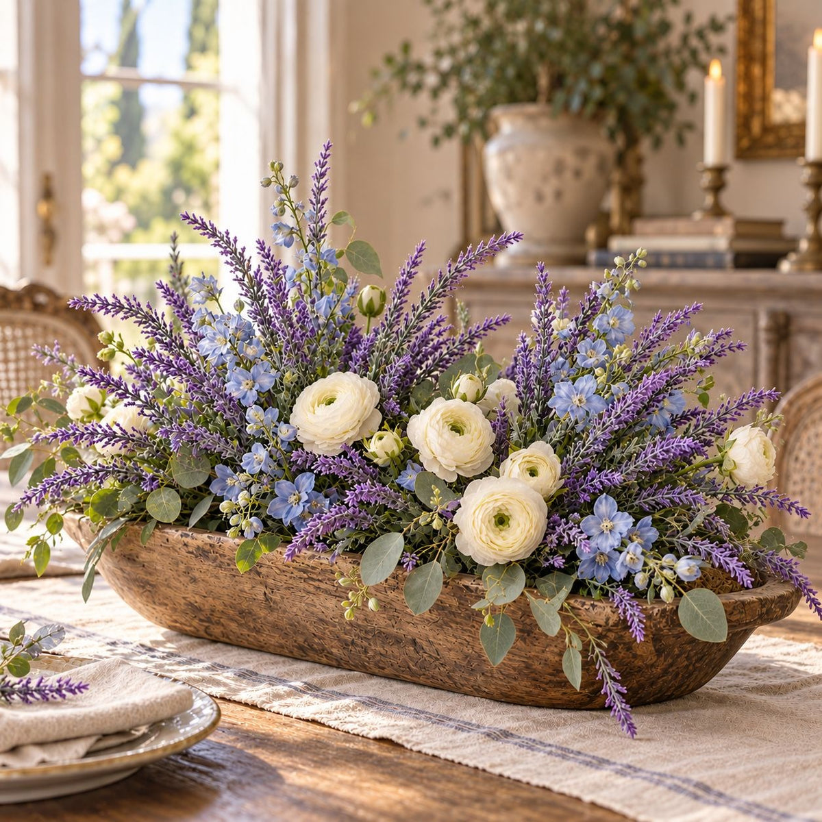 A rustic wooden bowl filled with a floral centerpiece arrangement of purple lavender, light blue flowers, cream ranunculus, and green eucalyptus, displayed on a dining table in a warm, sunlit room.