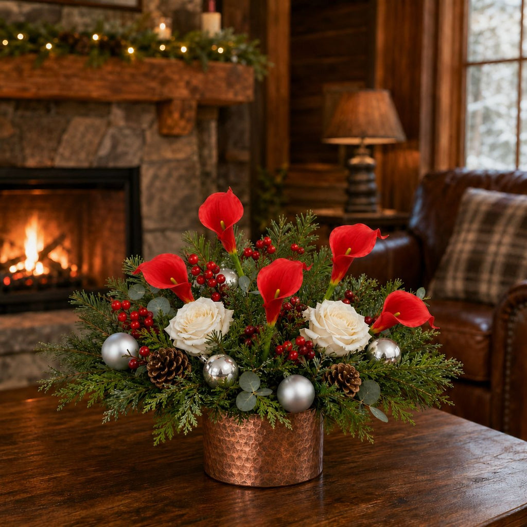 A festive holiday floral arrangement in a hammered copper pot, featuring red calla lilies, white roses, pinecones, silver ornaments, and green foliage, displayed on a wooden table in a cozy living room with a fireplace in the background.