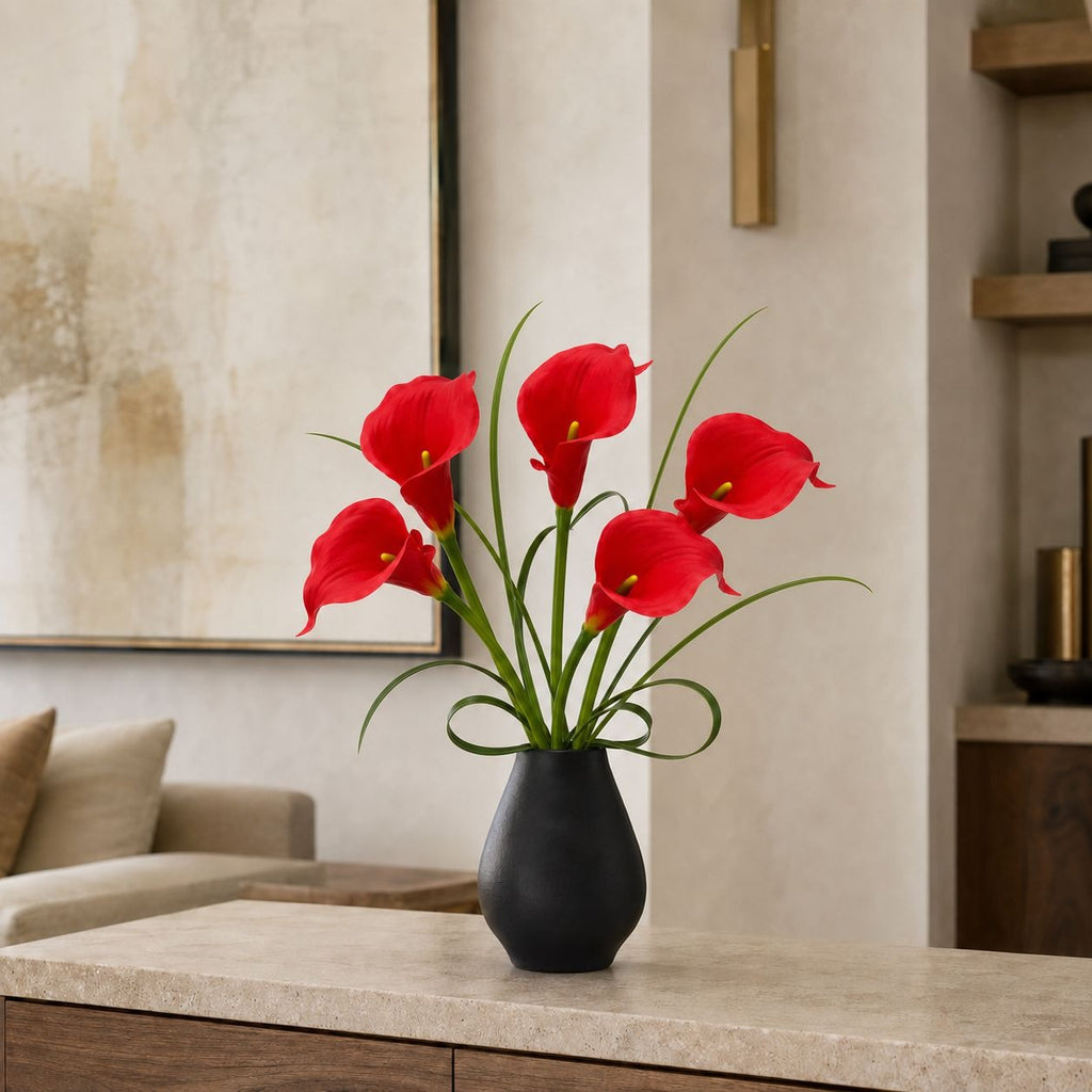 A black ceramic vase filled with bright red calla lilies and long green stems, placed on a light stone console table in a modern living room setting.