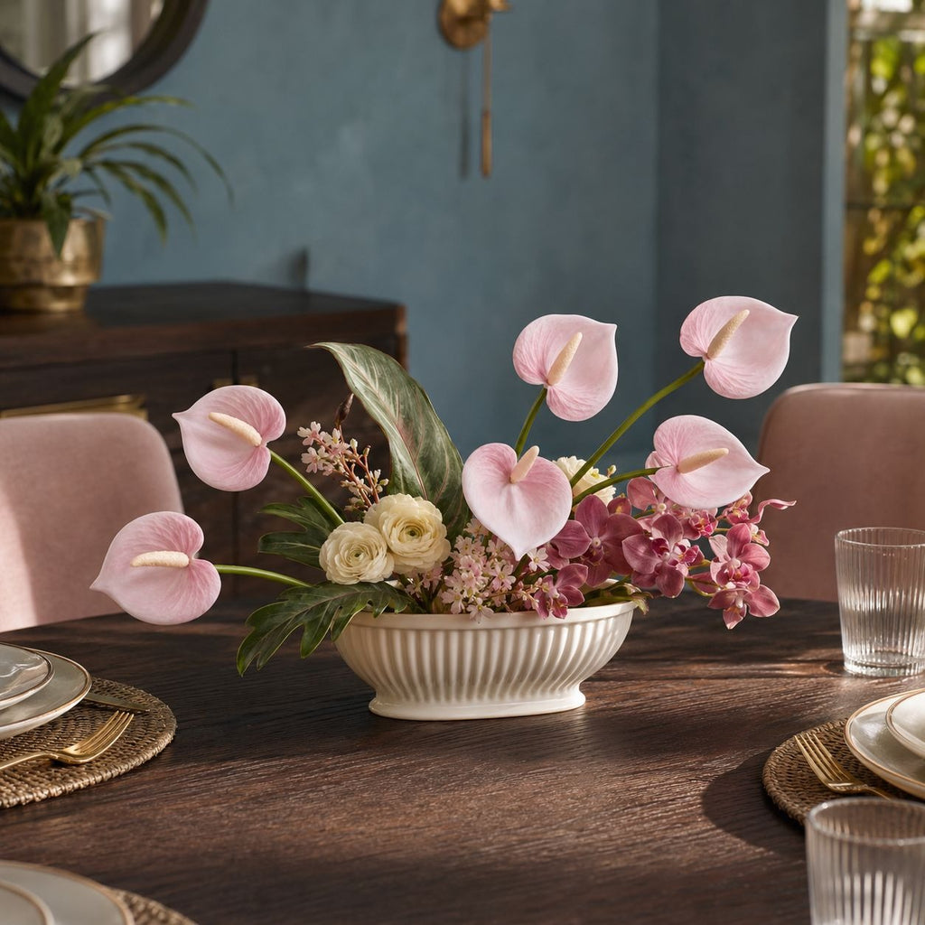A floral centerpiece in a ribbed white ceramic bowl, featuring pink anthuriums, pink orchids, cream roses, and green foliage, displayed on a dark wood dining table in a styled room.