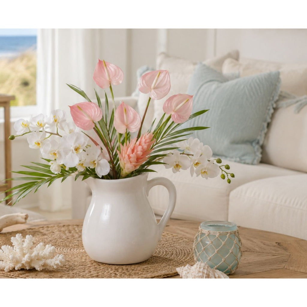 A white ceramic pitcher vase filled with a floral arrangement of pink anthuriums, white orchids, and green palm leaves, styled on a wooden table in a bright, modern living room.