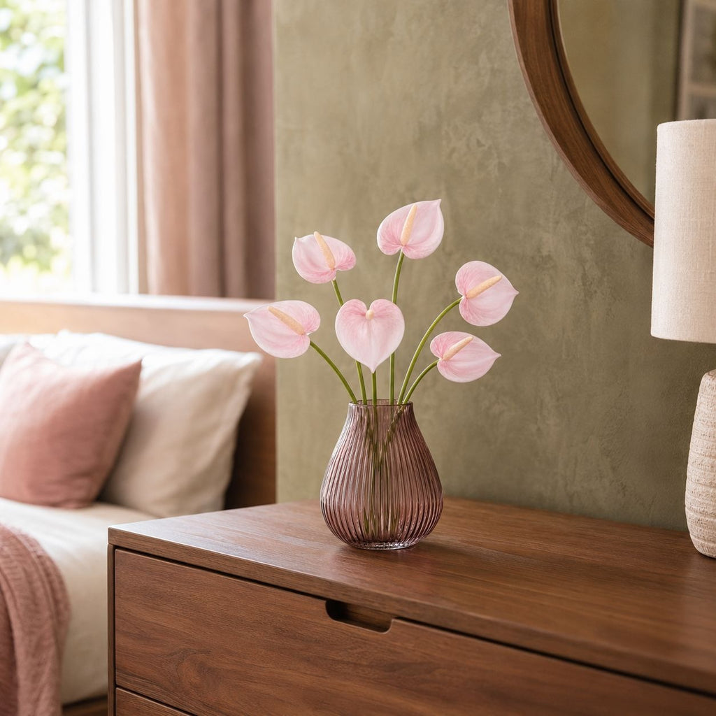 Pink anthurium flowers arranged in a ribbed mauve glass vase, placed on a wooden dresser in a softly lit bedroom setting.