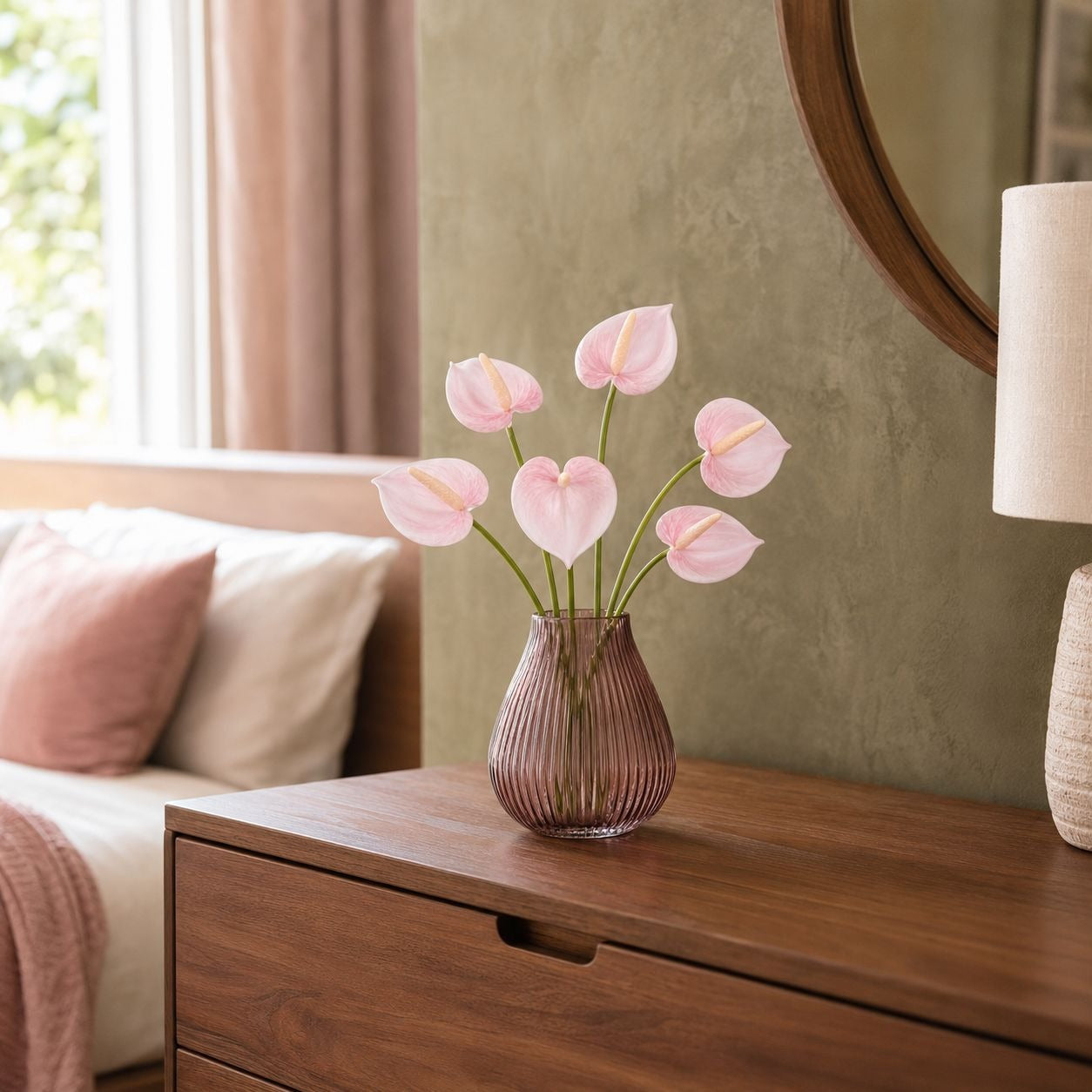 Pink anthurium flowers arranged in a ribbed mauve glass vase, placed on a wooden dresser in a softly lit bedroom setting.
