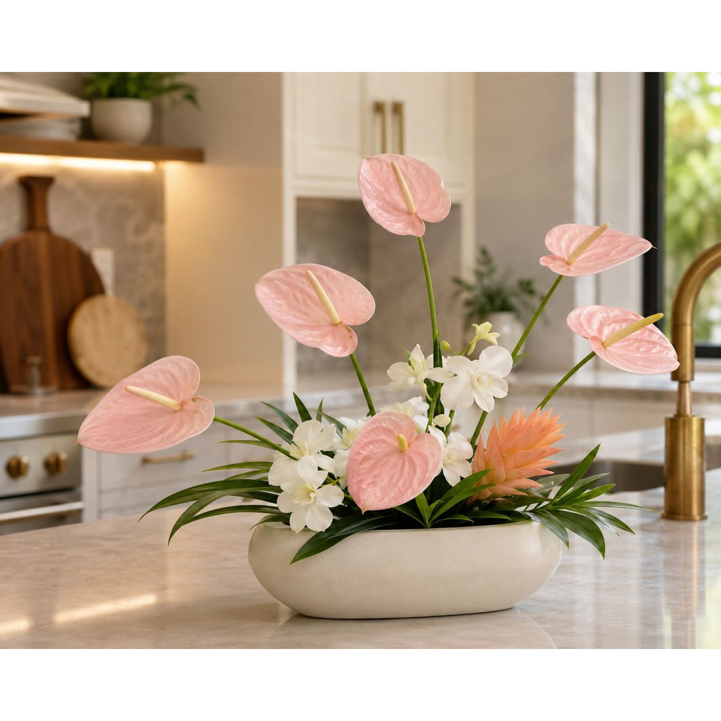 A floral arrangement of pink anthuriums, white flowers, and a peach pineapple in a round white ceramic pot, displayed on a marble kitchen countertop.