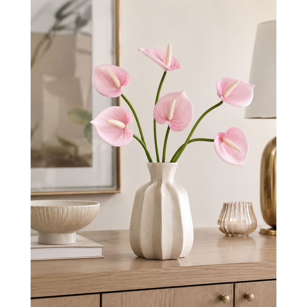 Pink anthurium flowers arranged in a ribbed off-white ceramic vase on a wooden console table, with a lamp, bowl, and framed art in the softly lit room background.