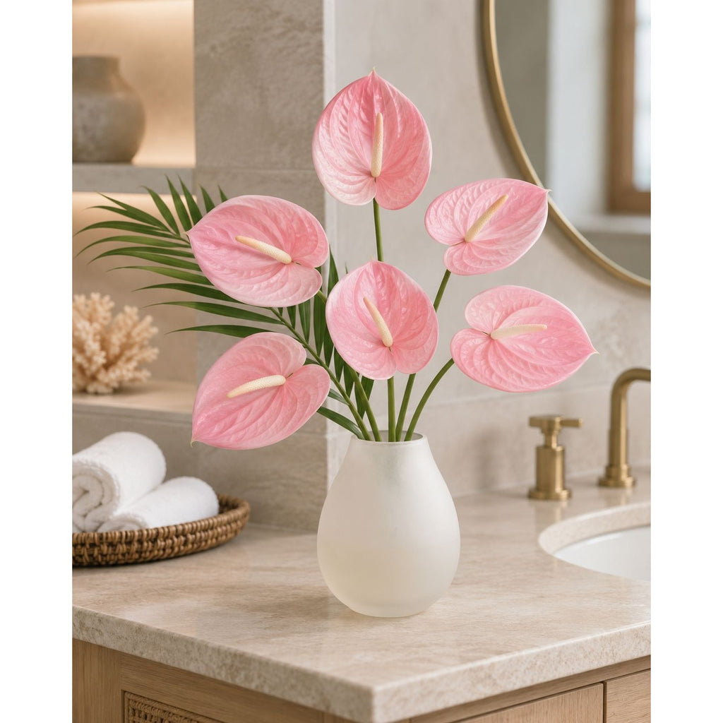 Six pink anthuriums in a white ceramic vase on a bathroom vanity next to a sink and mirror.