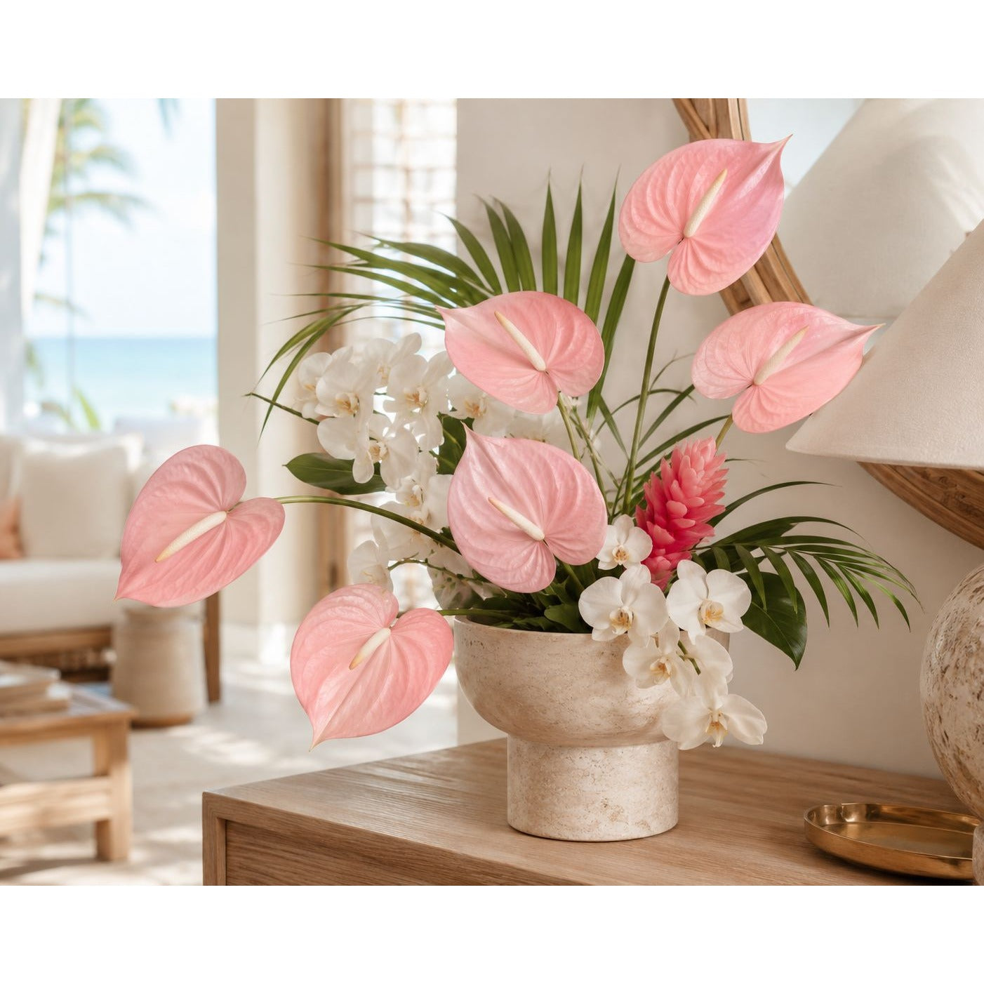 A floral arrangement in a textured cream-colored pot, featuring pink anthuriums, white orchids, and green palm leaves, displayed on a wooden table in a bright, modern living room setting.