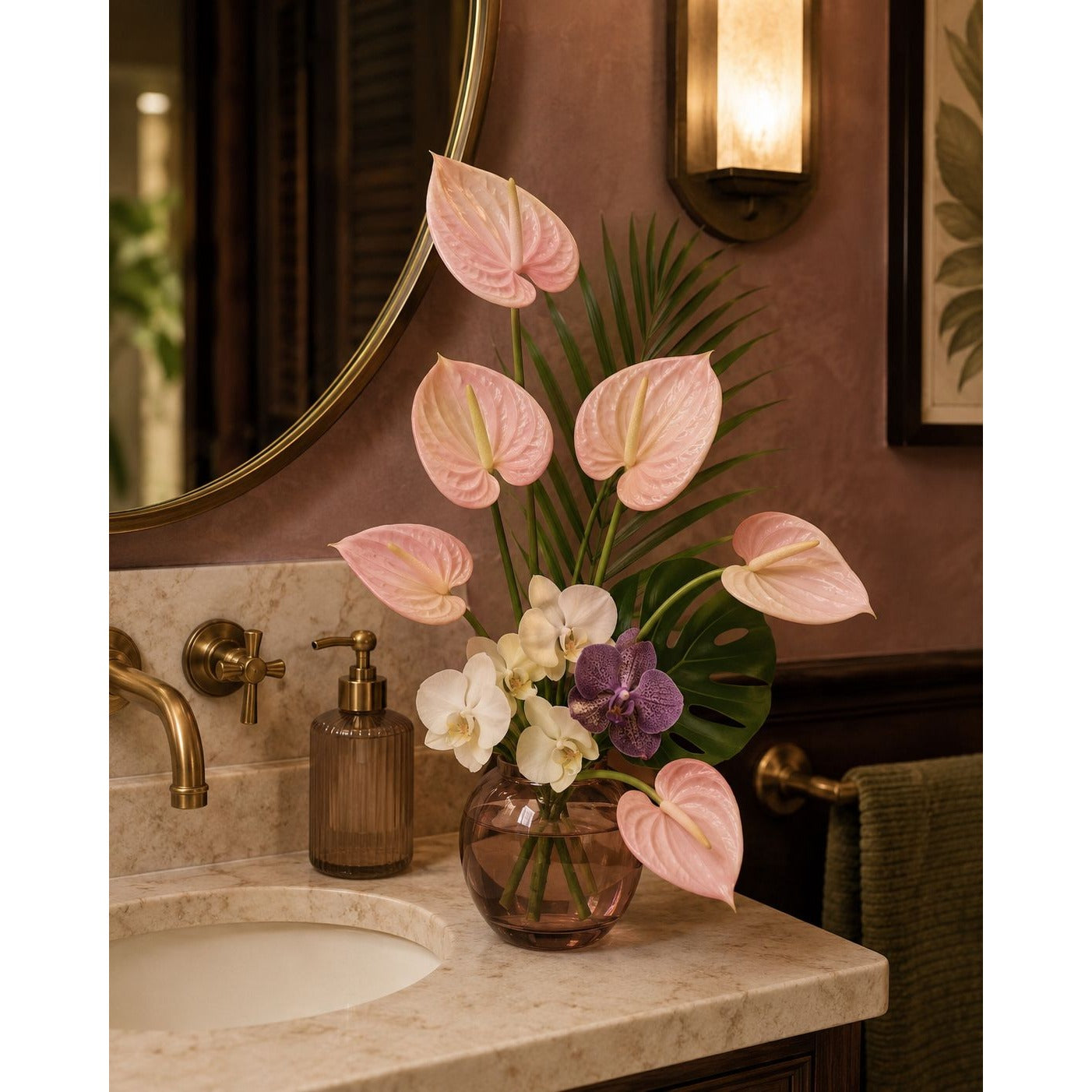 A floral arrangement of pink anthuriums, white orchids, and a purple flower in a round glass vase, displayed on a bathroom countertop beside a sink and brass fixtures.