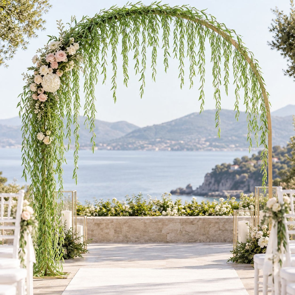 A romantic outdoor wedding arch setup with a large wooden arched structure draped in green leafy vines and decorated with white and blush pink flowers. A white aisle runner leads down to white chairs, with a scenic coastal bay and mountains in the background.
