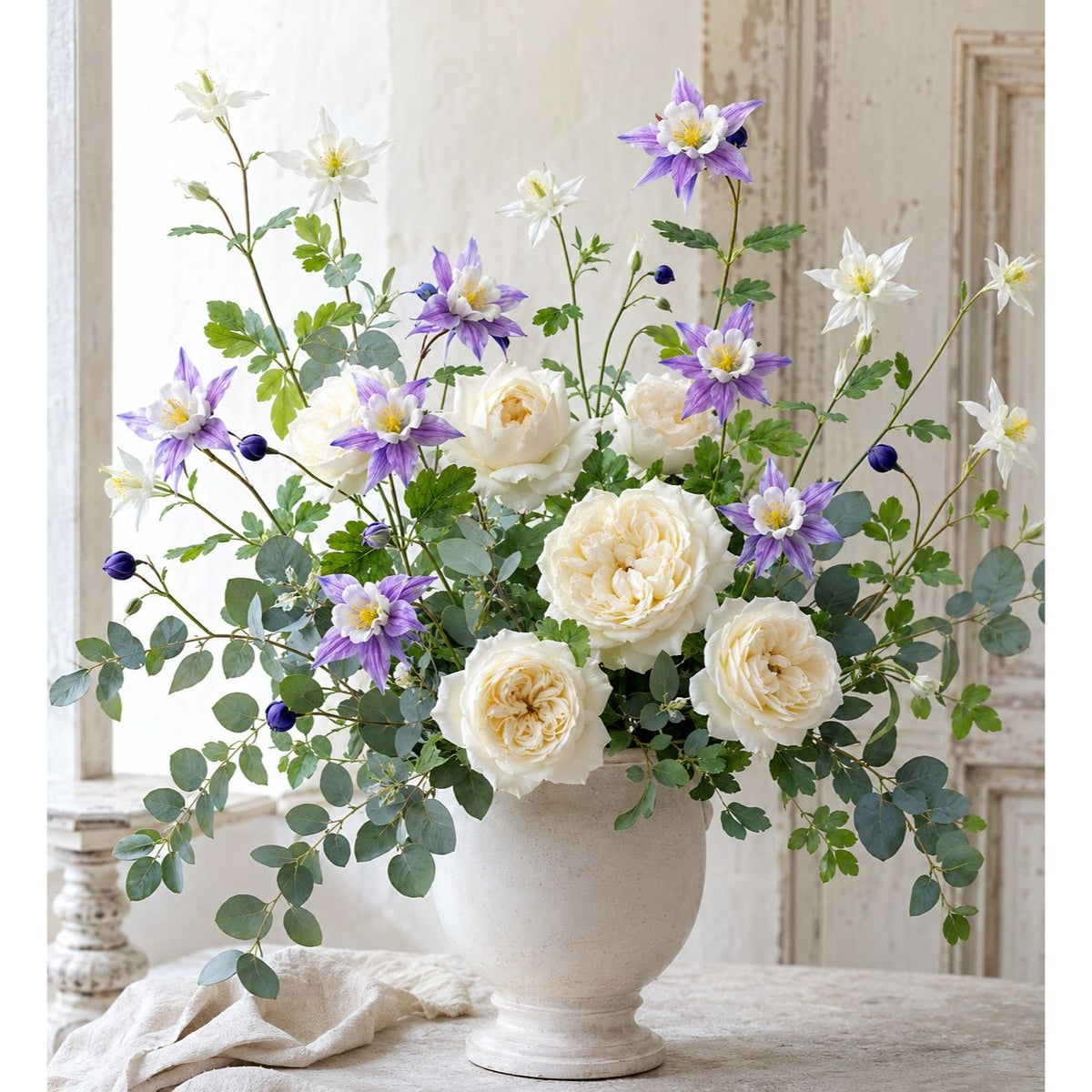 A large floral arrangement in a white pedestal vase, featuring cream roses, purple clematis, white starflowers, and green foliage, displayed on a table in a softly lit interior setting.