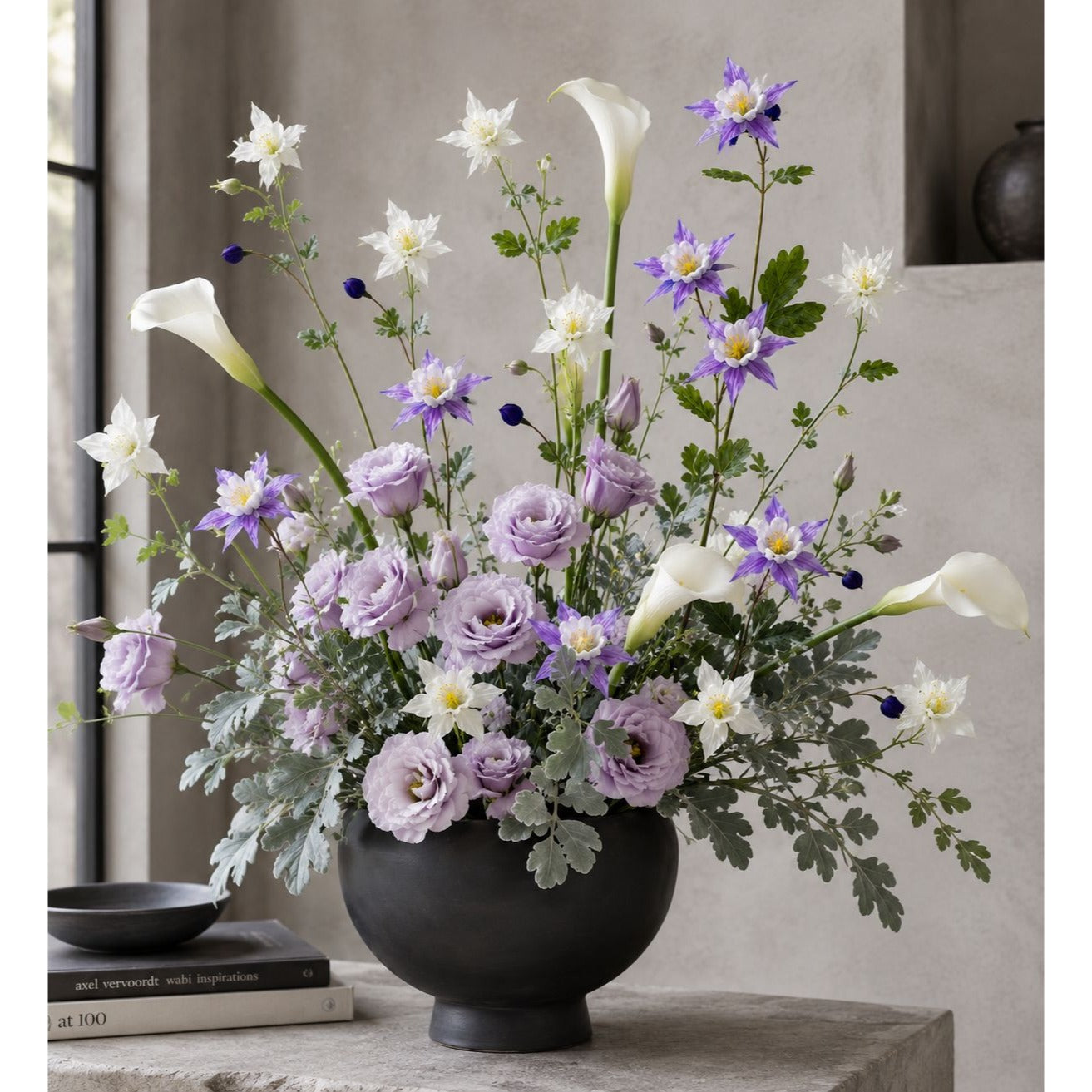 A large floral arrangement in a matte black round vase, featuring lavender and white flowers with green foliage, displayed on a stone table in a modern interior setting.