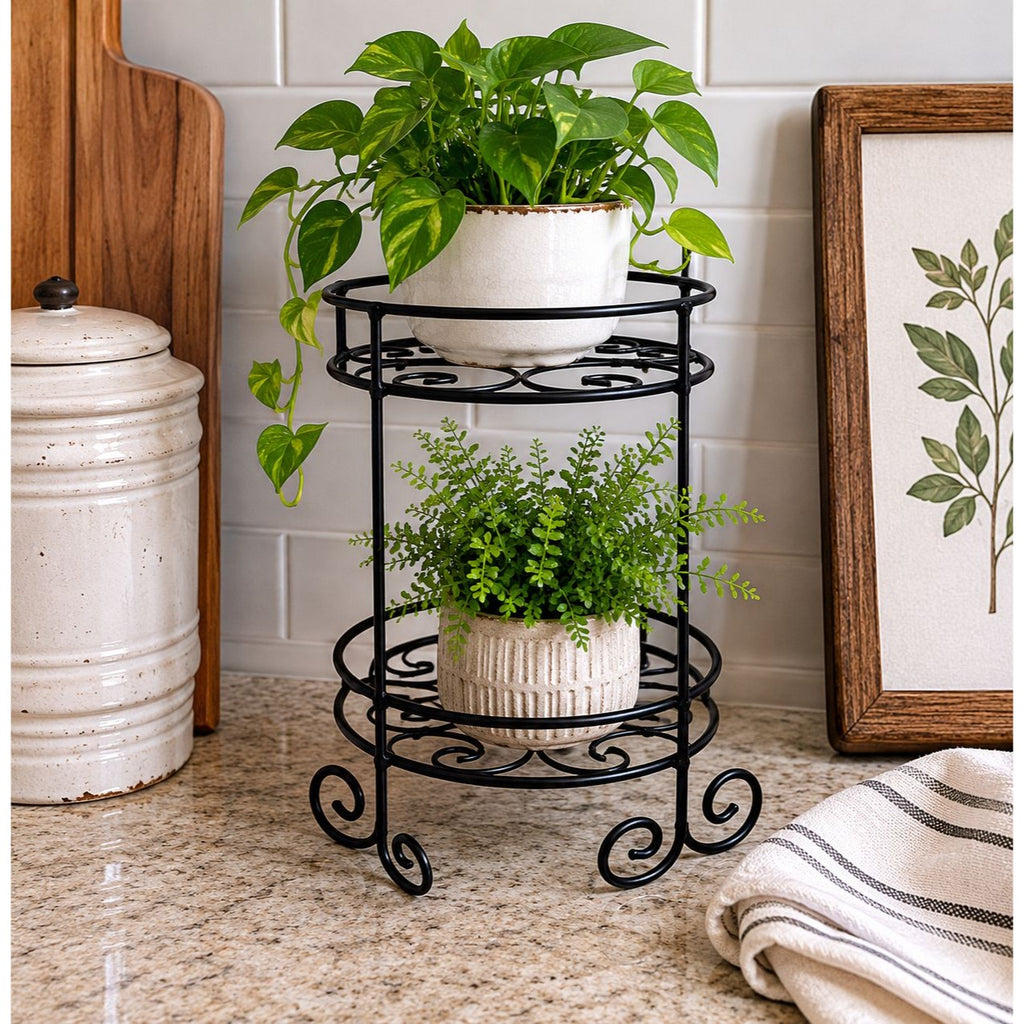 A two-tier black metal plant stand with scrollwork details, holding a green pothos plant in a white pot on the top shelf and a smaller green plant in a white pot on the bottom shelf, set on a kitchen countertop.