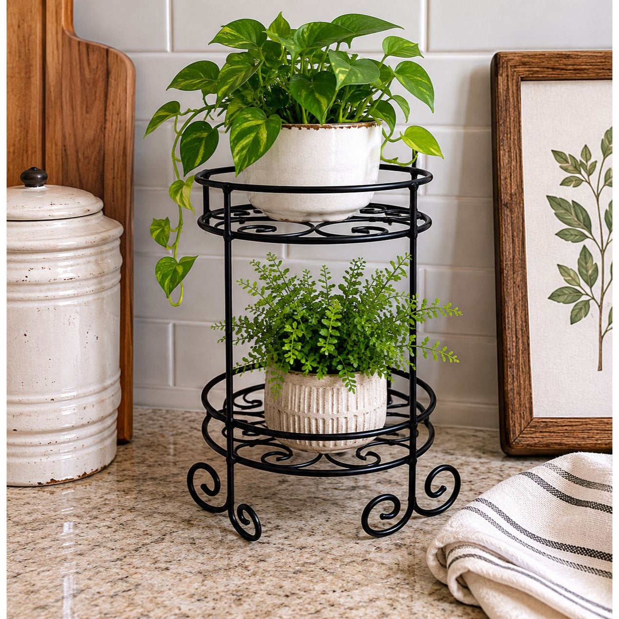 A two-tier black metal plant stand with scrollwork details, holding a green pothos plant in a white pot on the top shelf and a smaller green plant in a white pot on the bottom shelf, set on a kitchen countertop.