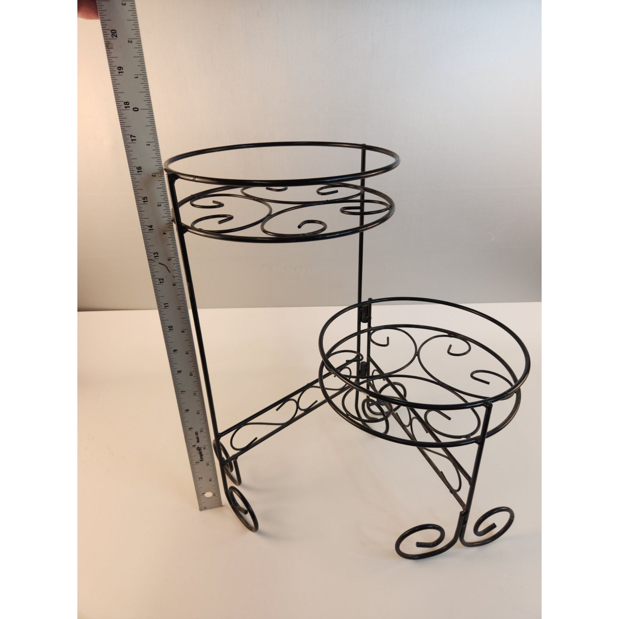 A black wrought iron double-tier plant stand with decorative scrollwork, shown next to a metal ruler to indicate height on a plain white background.