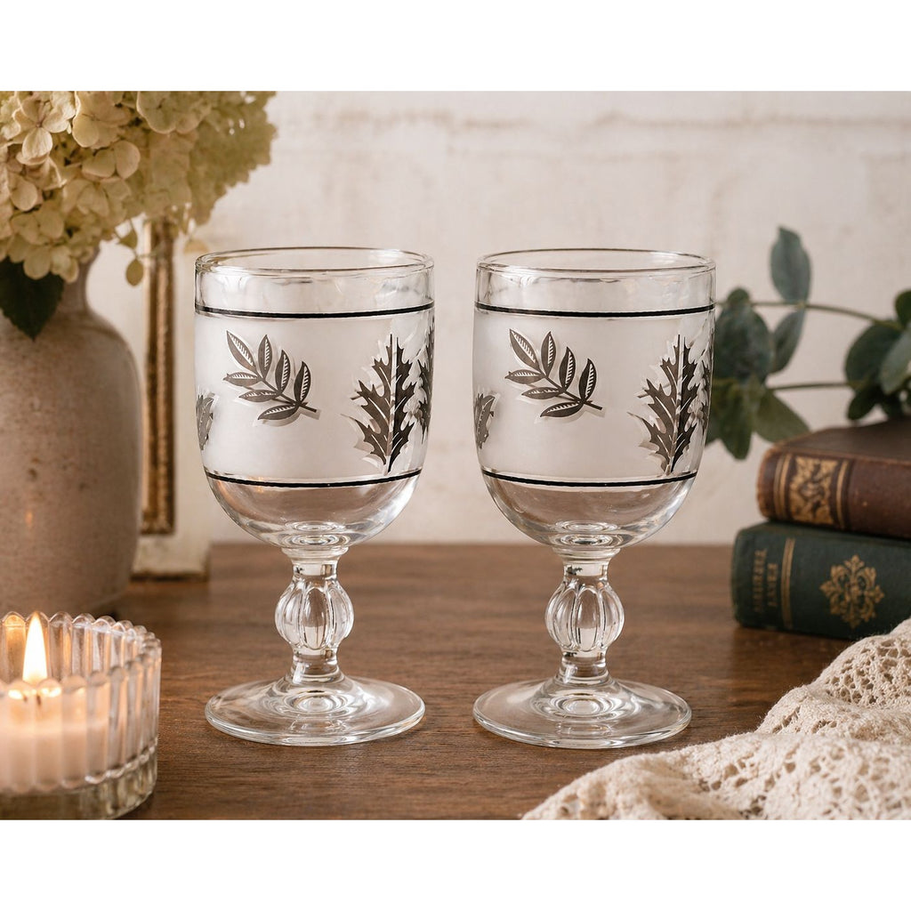 Two decorative glass goblets with leaf patterns on a wooden surface with books and flowers in the background.
