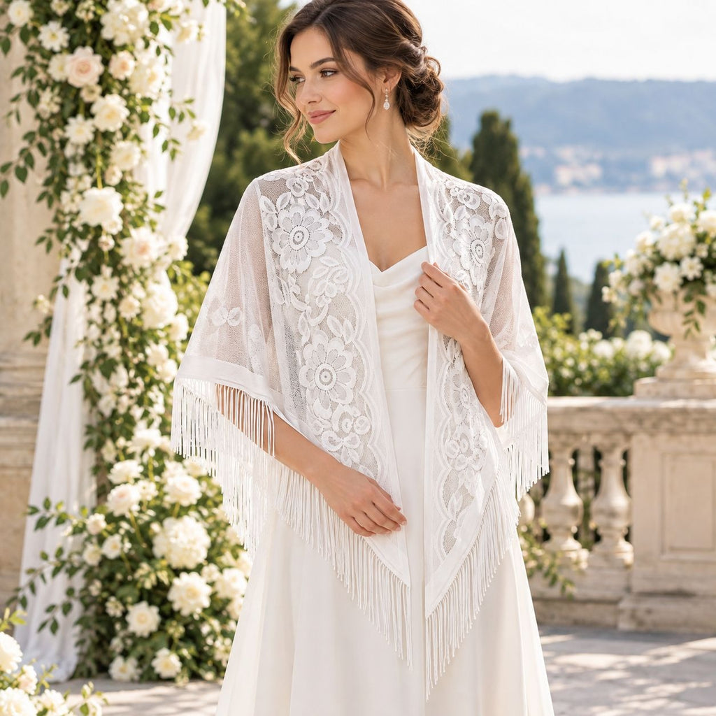 A woman wearing a white lace bridal shawl with fringe over a white wedding dress, standing in an outdoor garden setting with white rose arches and a scenic background.