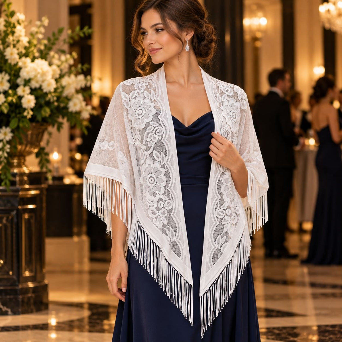 A woman in a formal indoor setting wears a white lace shawl with floral embroidery and fringe over a navy dress. The background shows a softly blurred elegant hall with flowers and guests.