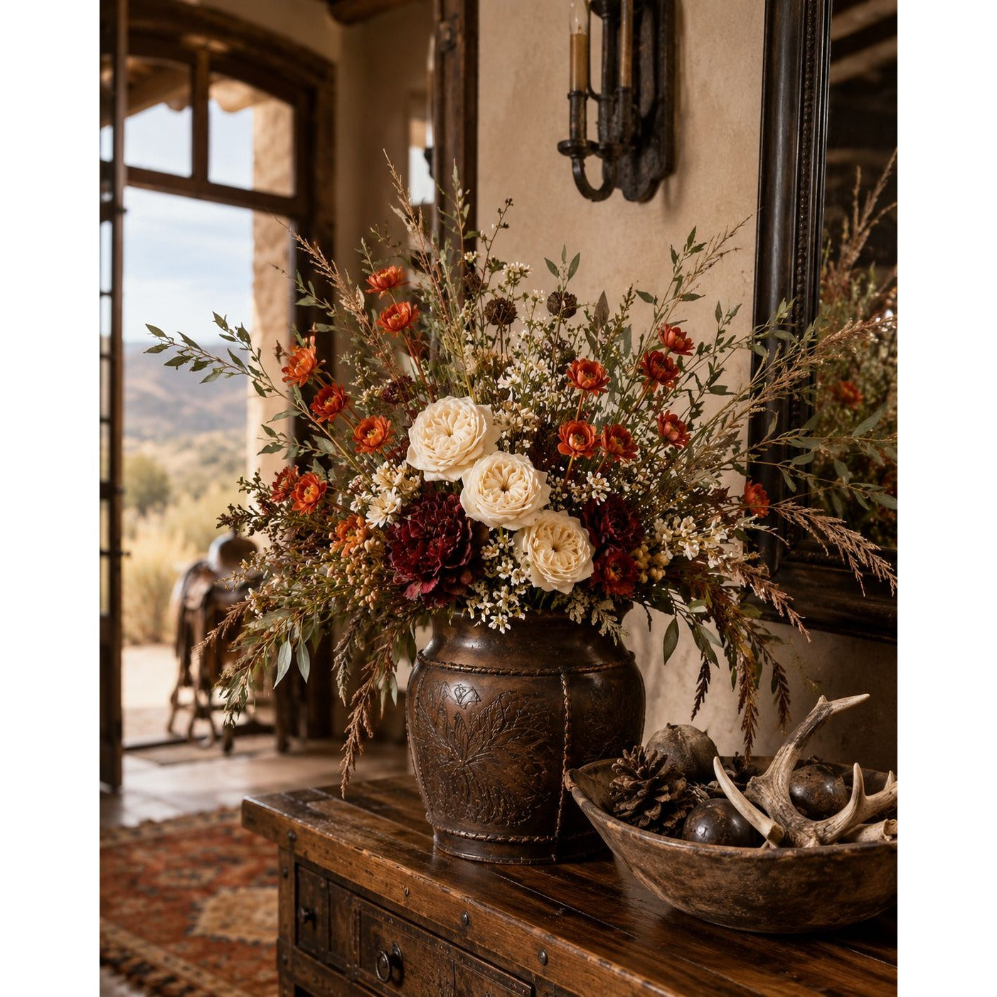 A large rustic ceramic pot filled with a dense arrangement of dried and fresh flowers, including cream roses, orange blossoms, burgundy dahlias, and green foliage, displayed on a wooden console table in a warm, rustic interior.