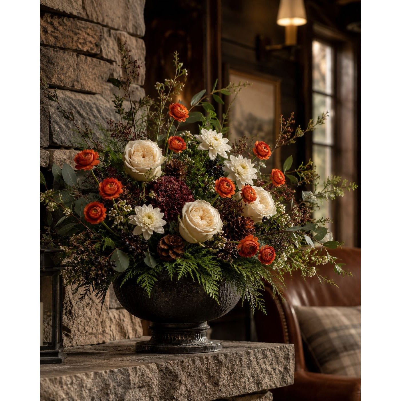 An autumn floral arrangement in a dark metal urn, featuring orange roses, cream dahlias, and green foliage, displayed on a stone mantel in a rustic indoor setting.
