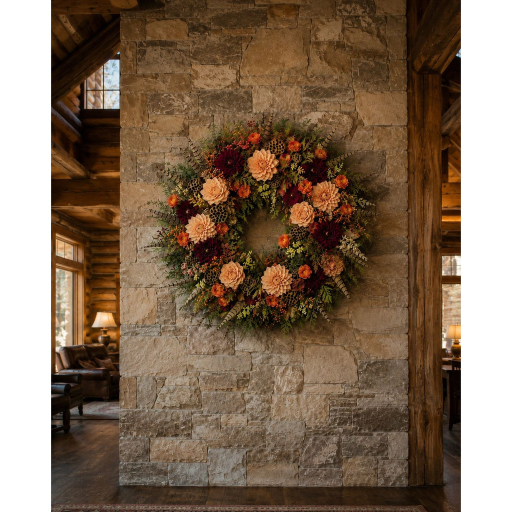 An autumn floral wreath with orange, burgundy, and cream flowers, hung on a rustic stone pillar inside a cozy lodge-style room with wooden beams and a fireplace.