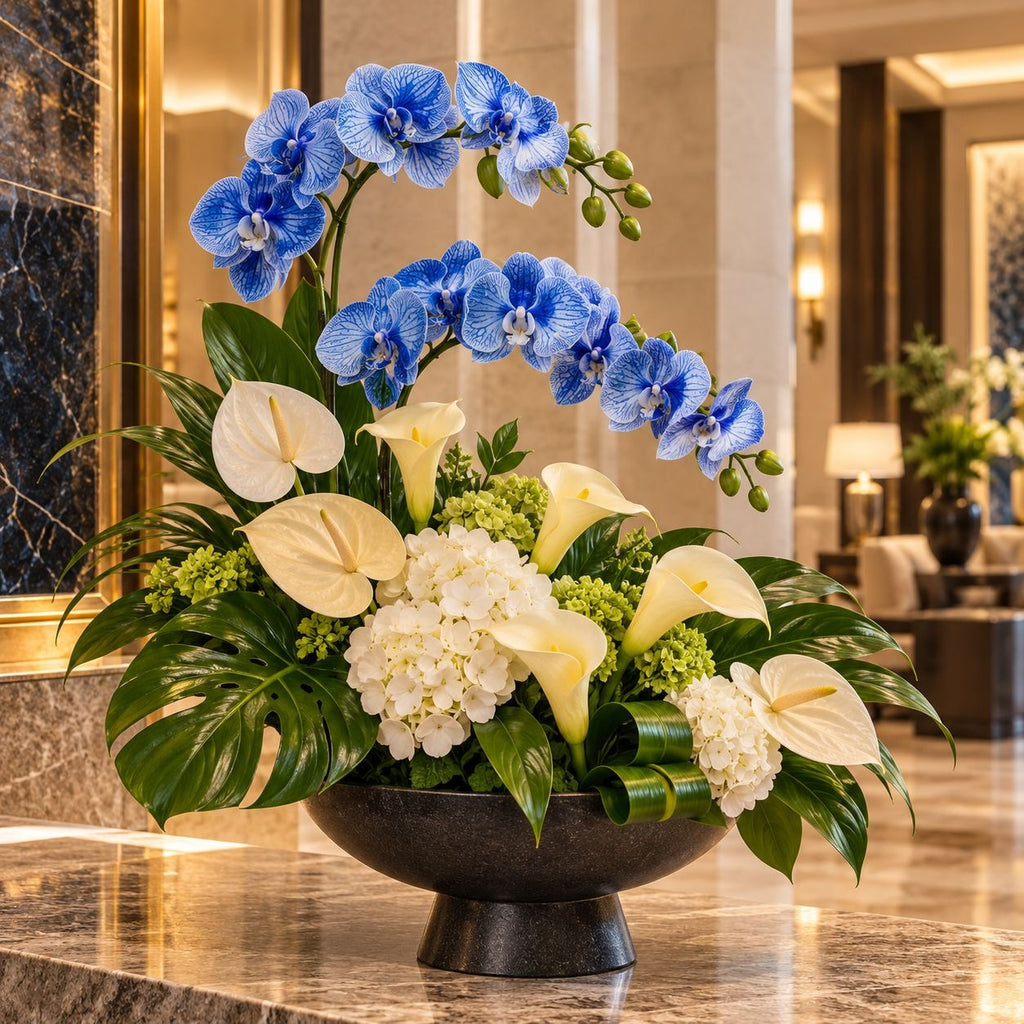 A large floral arrangement in a dark round pedestal bowl, featuring blue orchids, white calla lilies, white anthuriums, green hydrangeas, and green foliage, displayed on a polished marble counter in a luxury lobby.