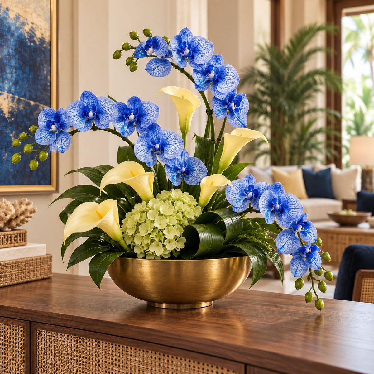 A vibrant floral arrangement featuring blue orchids, yellow calla lilies, and light green hydrangeas in a polished gold bowl, displayed on a wooden console table in a modern living room setting.