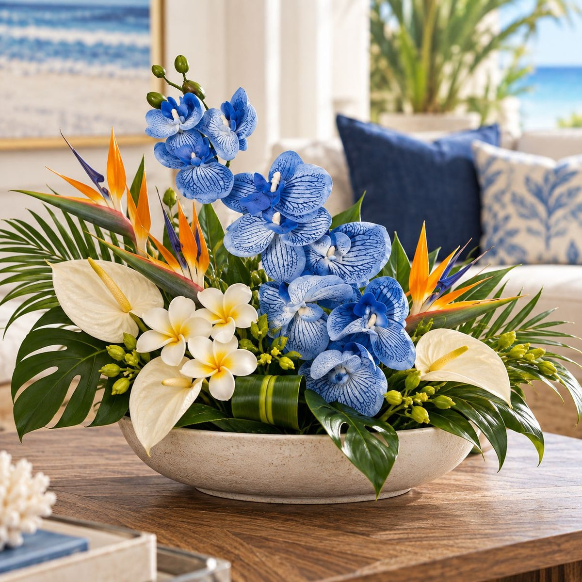A tropical floral arrangement in a round beige bowl, featuring blue orchids, white plumeria, orange bird of paradise, green palm leaves, and white anthuriums, displayed on a wooden table in a bright living room setting.